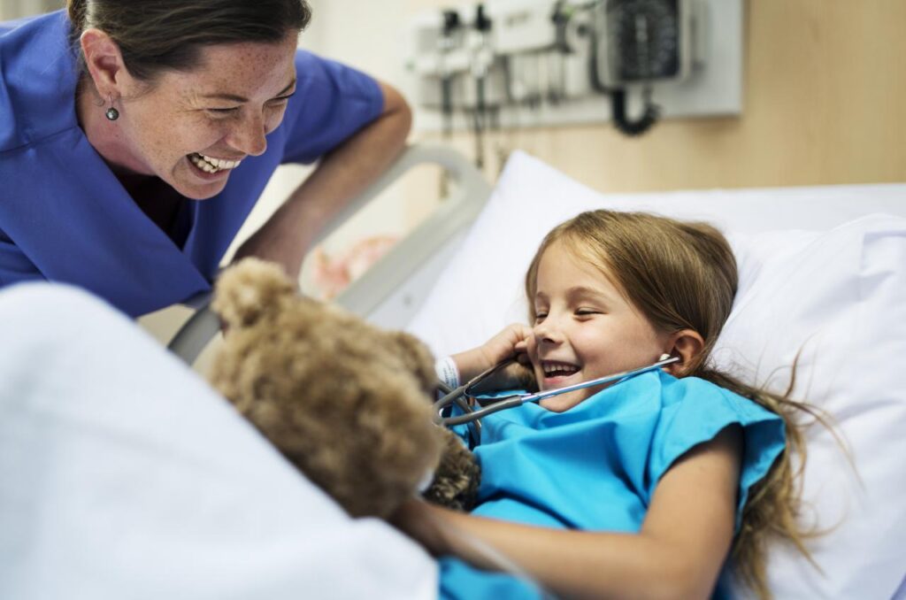 A female nurse laughs with her child patient as they play with a teddy bear