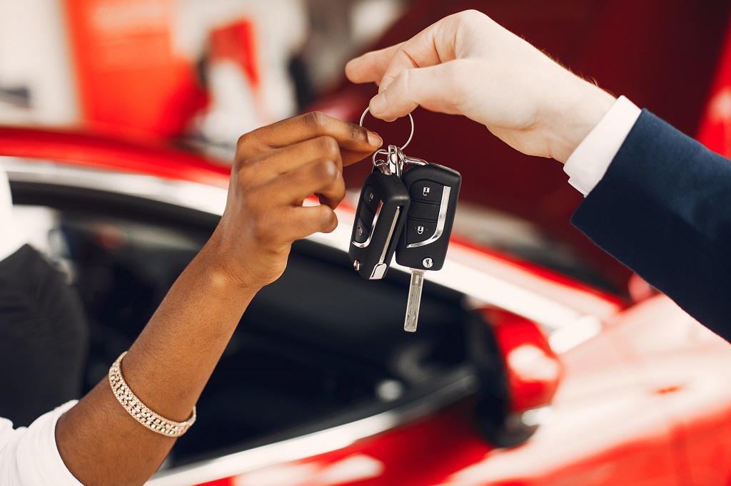 A car dealer hands car keys to a new buyer sat in a red car. 