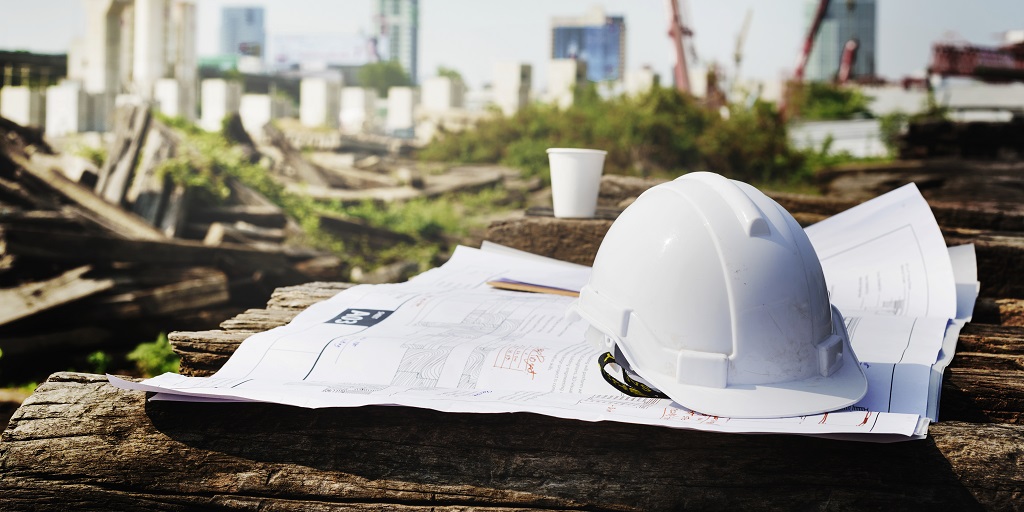 A hard hat and some blueprints on the ground in front of a construction site.